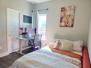 Een slaapkamer met een bed, een bureau met een computer, een stoel, een raam met gordijnen en een schilderij aan de muur.