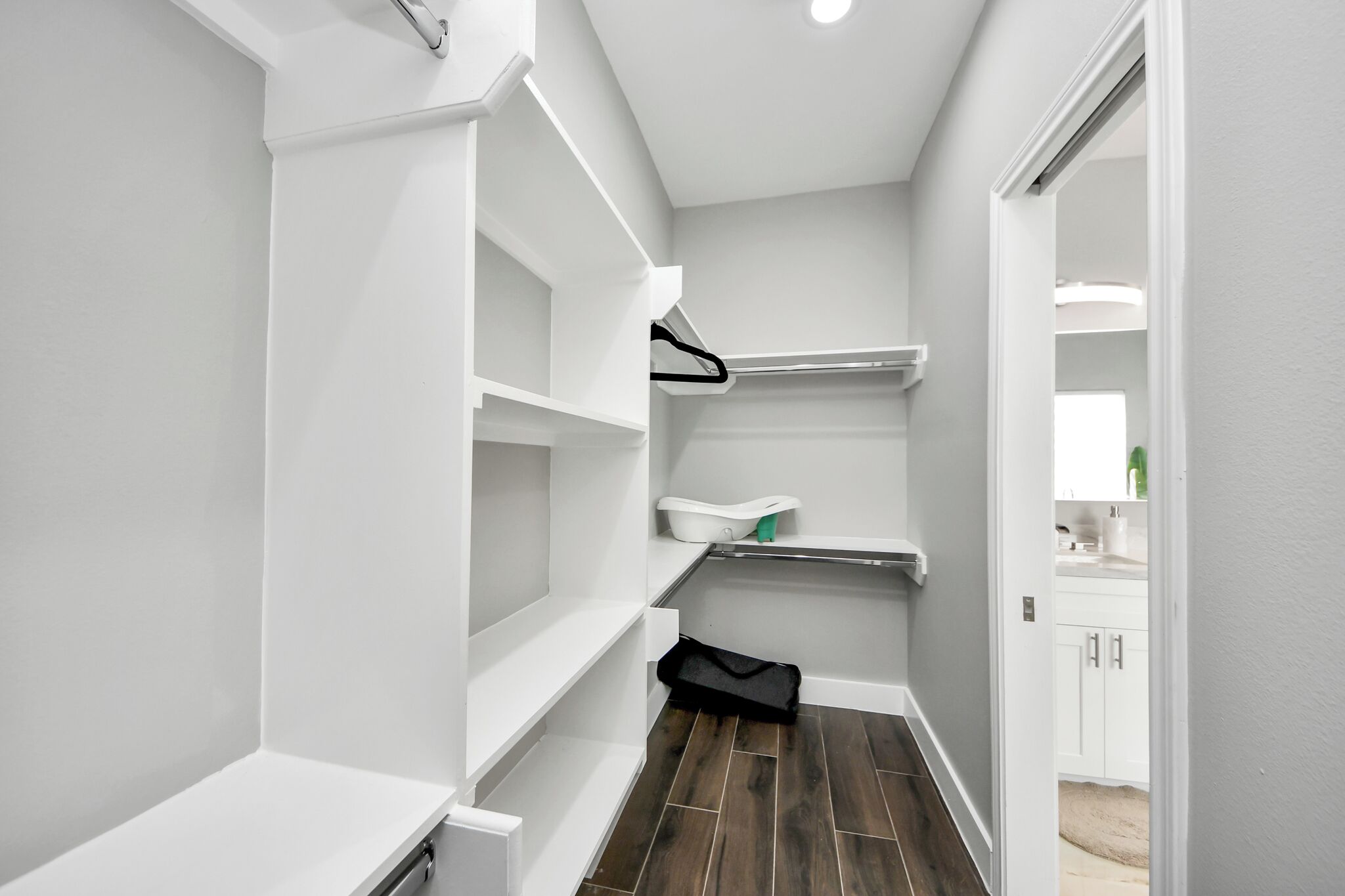 A narrow hallway with white shelving units, a black bag on the floor, and a door leading to a bathroom with a mirror and sink.