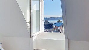 Une pièce dotée d’une grande fenêtre, d’un mur blanc et offrant une vue sur une zone côtière avec des bâtiments et une étendue d’eau.