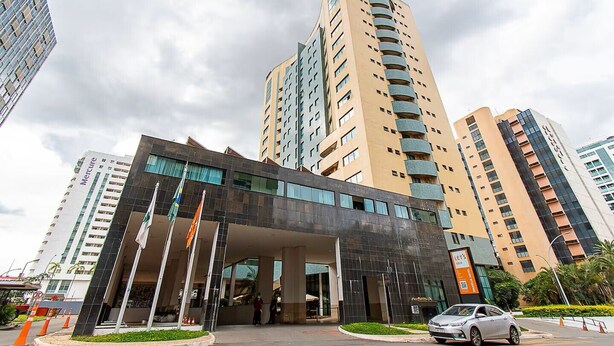 A modern hotel entrance with a glass facade, multiple floors, and a parking area with a car. - Flat Let's Idea - Centro de Brasília (Brasília)