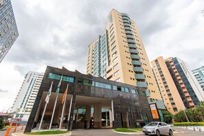 A modern hotel entrance with a glass facade, multiple floors, and a parking area with a car. - Flat Let's Idea - Centro de Brasília (Brasília)