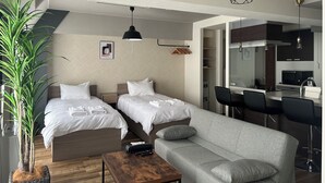 A modern hotel room with two beds, a grey sofa, a wooden coffee table, a large potted plant, and a kitchen area visible through an open door. - TOWN ROYAL 701 (Sapporo)