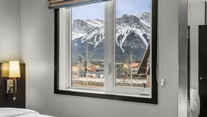 A hotel room with a bed, a window with a mountain view, a lamp, and a door. - Mountain Views | Resort Pool w/Waterslide, Hot Tub (Canmore)