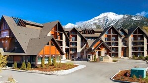 A lodge with a symmetrical, multi-story design, featuring a central courtyard and a mountain backdrop. - Mountain Views | Resort Pool w/Waterslide, Hot Tub (Canmore)