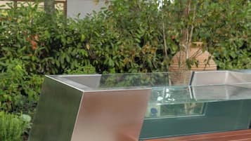 Una fuente moderna de metal con superficie reflectante, rodeada de abundante vegetación.