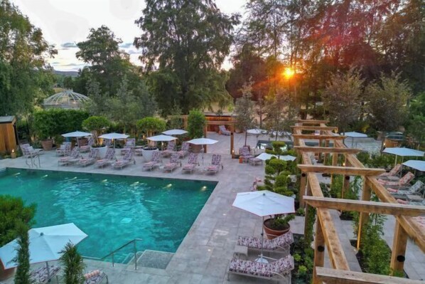 A pool area with lounge chairs and umbrellas, surrounded by trees and a wooden pergola.