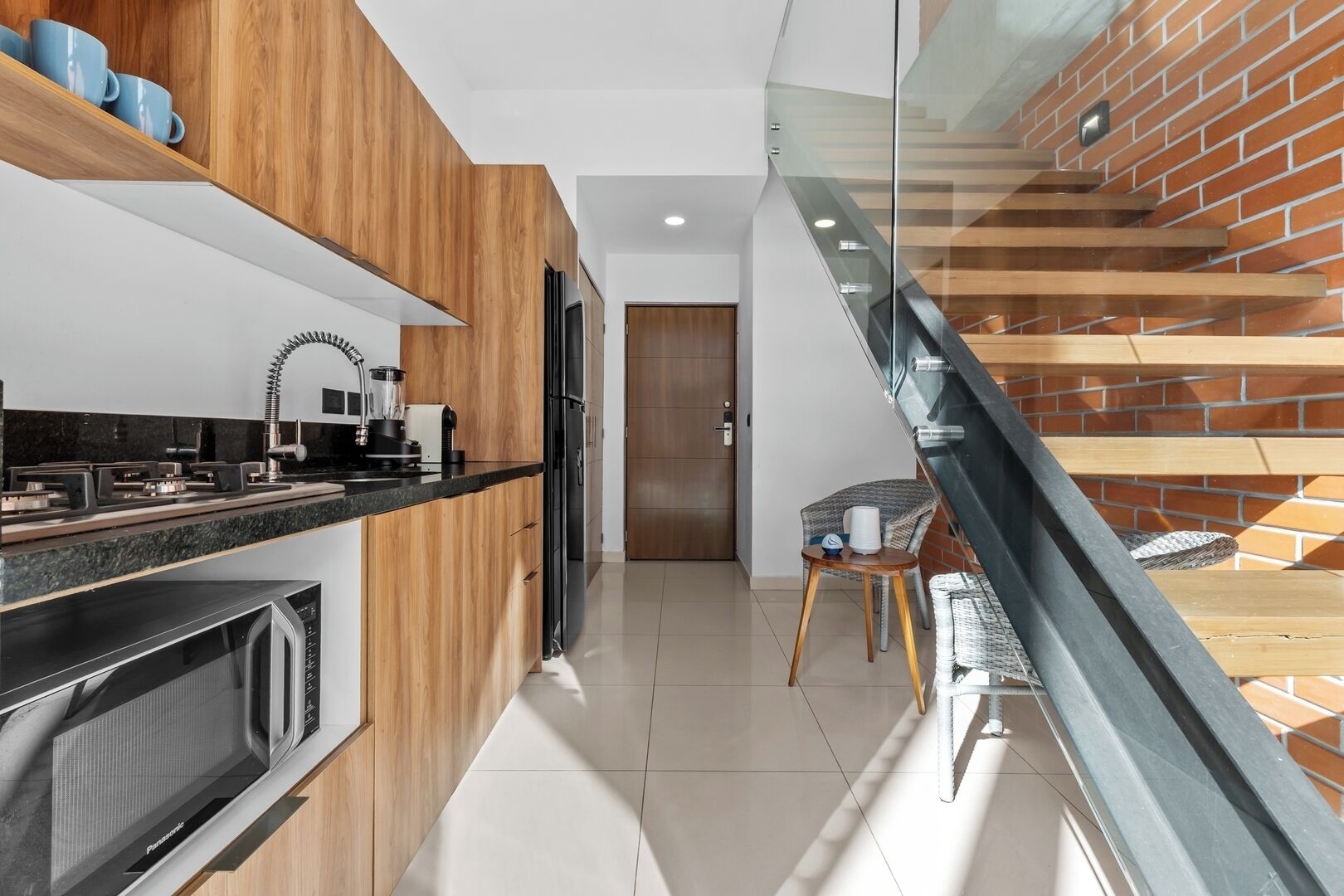 Una cucina moderna con mobili in legno, un piano di lavoro nero e un forno a microonde integrato. Si vede una scala in vetro che conduce a un livello inferiore.