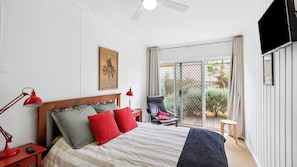 Une chambre à coucher avec un lit, deux tables de chevet avec des lampes, une chaise, un petit tabouret, une télévision et un balcon donnant sur un espace verdoyant.