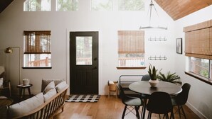 Ein gemütliches Interieur mit Holzbalkendecke, einem Essbereich mit rundem Tisch und Stühlen, einer Couch und einer Tür mit Blick ins Freie.