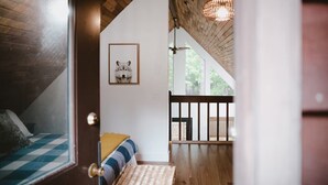 Ein gemütliches Zimmer mit Holzboden, einem gerahmten Bild an der Wand, einem Bett mit gestreifter Decke und Blick ins Freie durch ein Fenster.