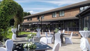 An outdoor patio with tables and chairs, a brick building in the background, and a large tree. - Delta Hotels by Marriott Peterborough (Orton Waterville)