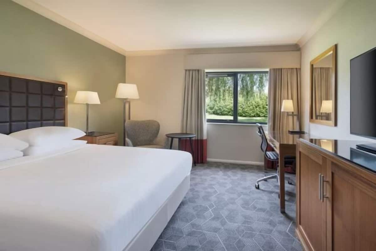 A hotel room with a large bed, two bedside lamps, a desk with a chair, a television, and a window with curtains.