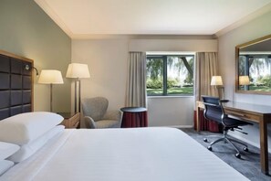 A hotel room with a bed, two chairs, a desk, a lamp, a mirror, and a window with a view of greenery. - Delta Hotels by Marriott Peterborough (Orton Waterville)
