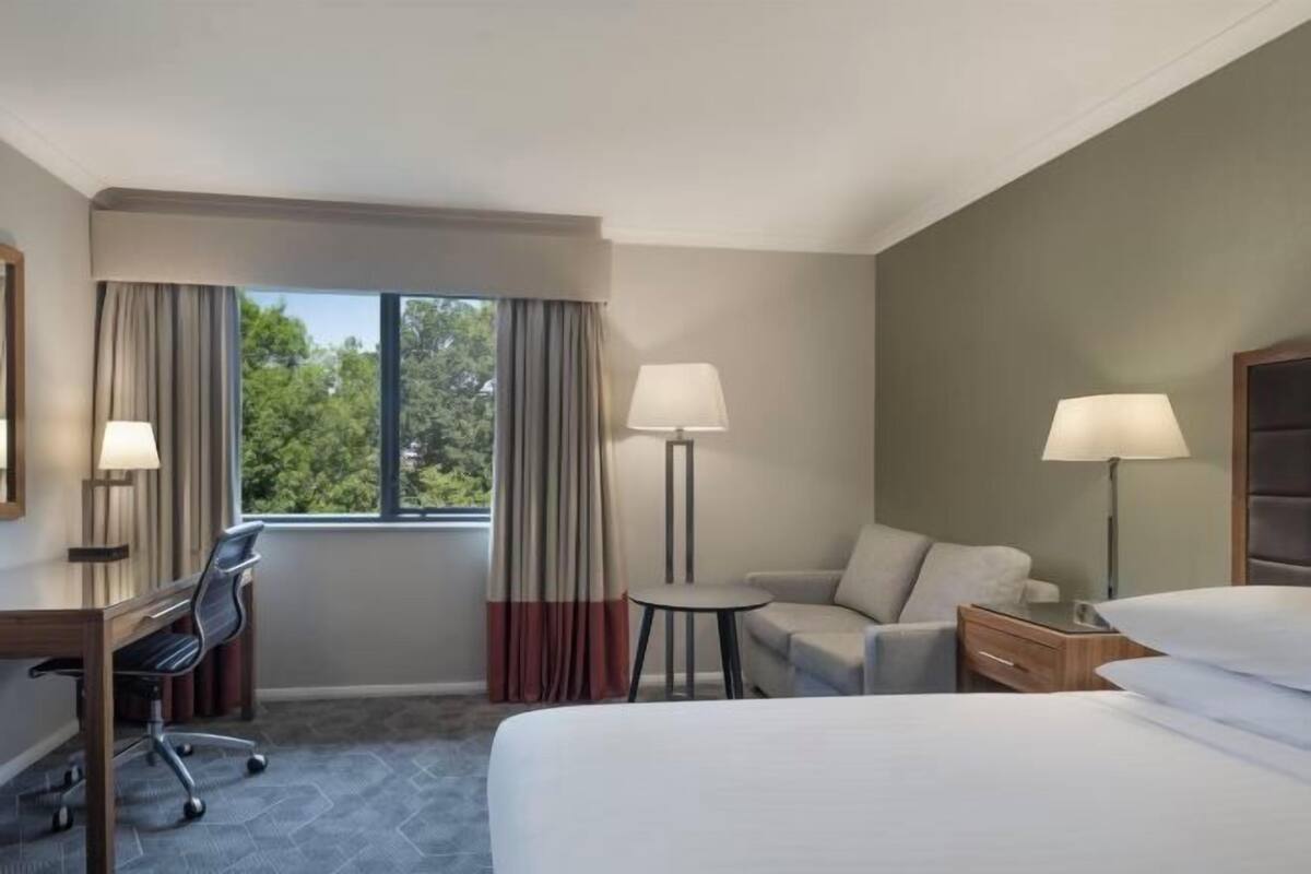 A hotel room with a bed, a desk, a chair, a sofa, a nightstand, and a window with curtains.