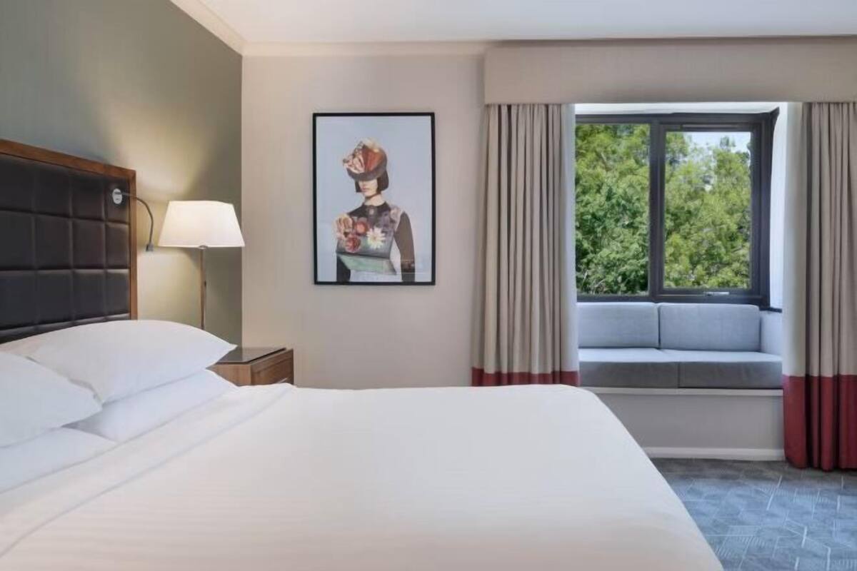 A hotel room with a bed, a nightstand, a lamp, a window with curtains, and a wall-mounted picture.