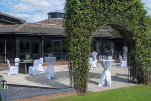 An outdoor patio with white chairs and tables, an archway with greenery, and a building with large windows in the background. - Delta Hotels by Marriott Peterborough (Orton Waterville)