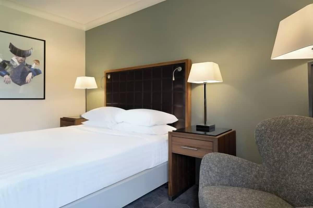 A hotel room with a bed, two bedside lamps, a nightstand, a chair, and a framed picture on the wall.