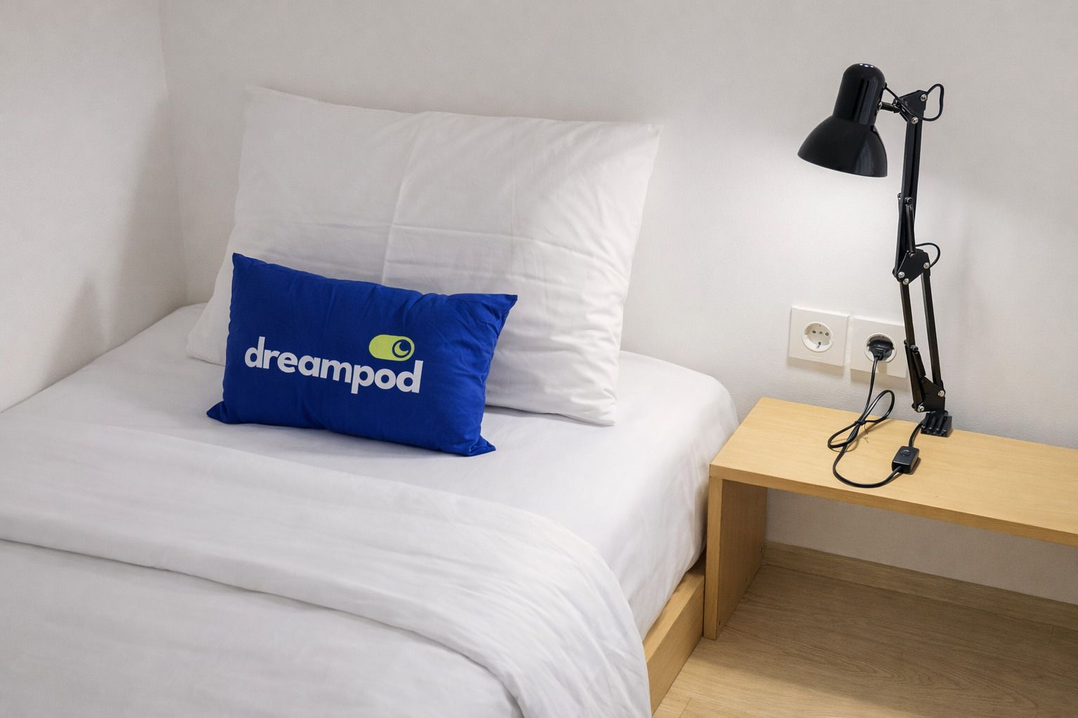 Ein Bett mit weißer Bettwäsche, ein blaues Kopfkissen mit dem Aufdruck 'dreampod' und eine schwarze Schreibtischlampe auf einem Holznachttisch.
