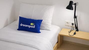 Ein Bett mit weißer Bettwäsche, ein blaues Kopfkissen mit dem Aufdruck 'dreampod' und eine schwarze Schreibtischlampe auf einem Holznachttisch.