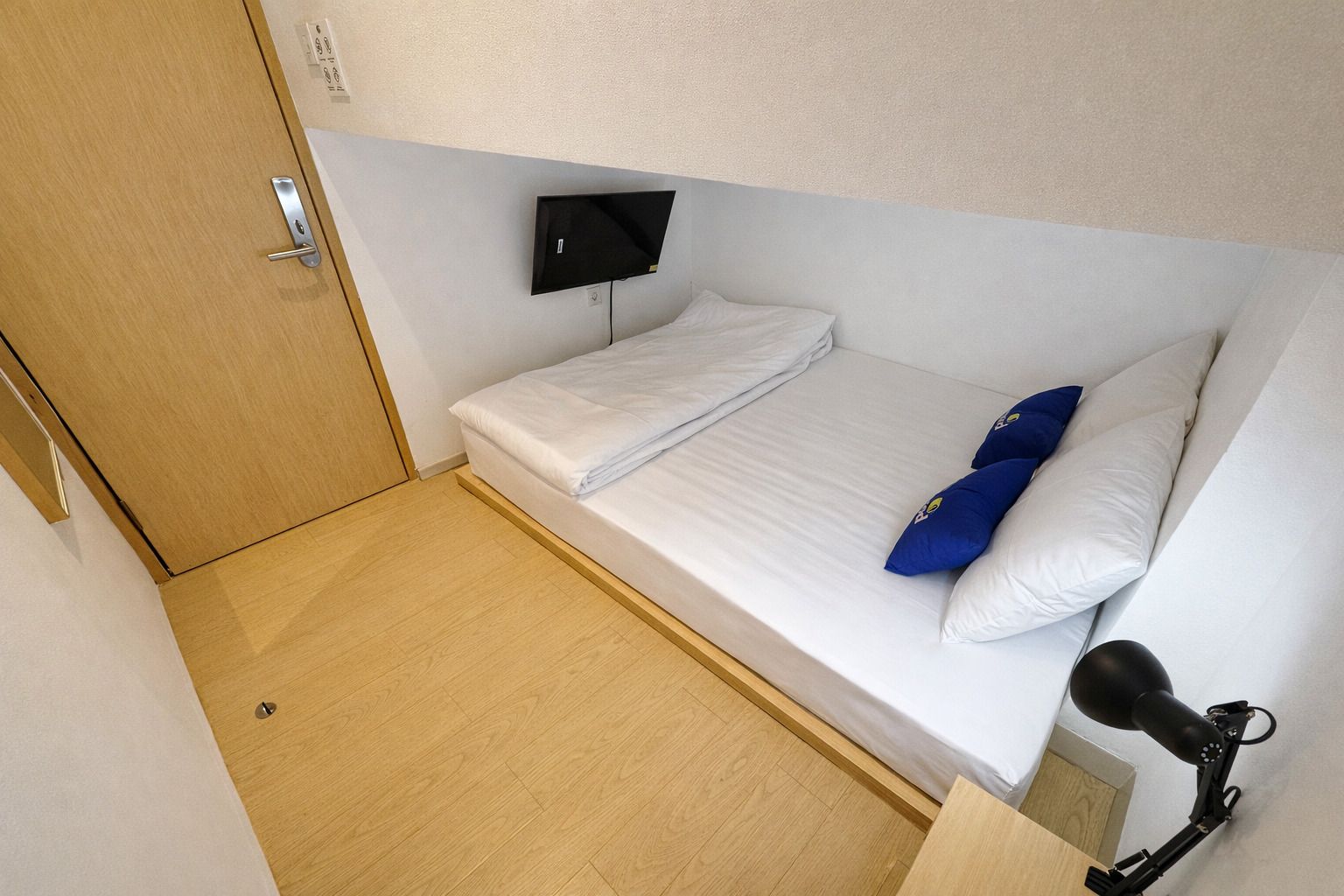 Una habitación pequeña con cama individual, piso de madera, televisor montado y un micrófono sobre un soporte.