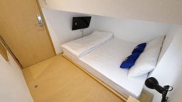 Una habitación pequeña con cama individual, piso de madera, televisor montado y un micrófono sobre un soporte.