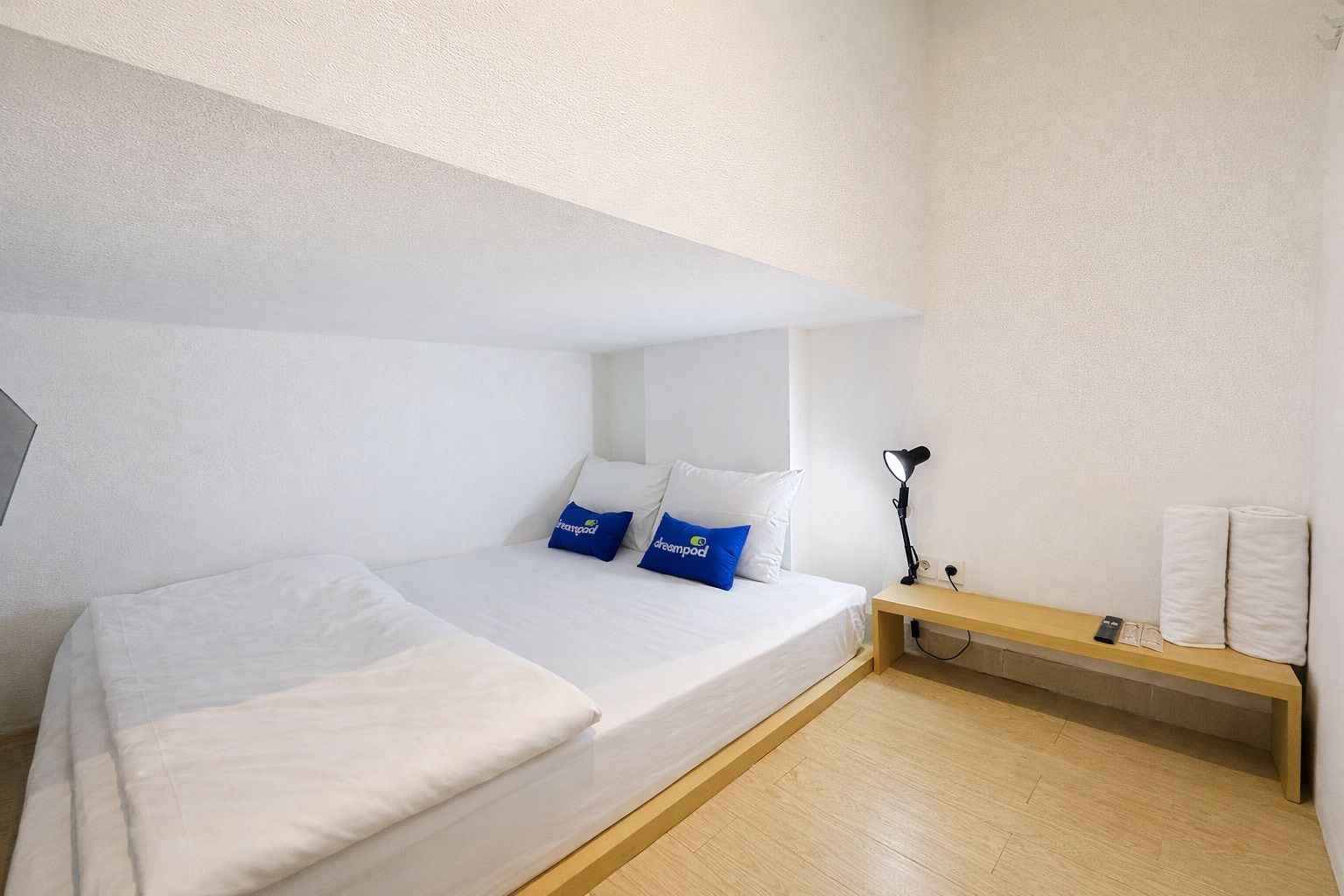 A minimalist hotel room with a single bed, a bedside lamp, a small wooden bench, and a wall-mounted shelf with towels.