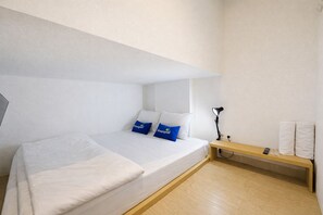 Een strakke hotelkamer met een eenpersoonsbed, een bedlampje, een klein houten bankje en een wandplank met handdoeken.
