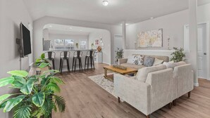 A modern living room with a sofa, coffee table, and bar stools. There is a kitchen area in the background with a large window. - Convenient Family Home in Heart of Conroe (Conroe)