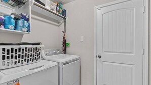 A laundry room with a washing machine, dryer, and shelves with detergent bottles and other supplies. - Convenient Family Home in Heart of Conroe (Conroe)