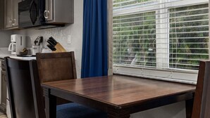 Una cocina con una mesa de comedor y sillas, microondas, cafetera y ventana con persianas.