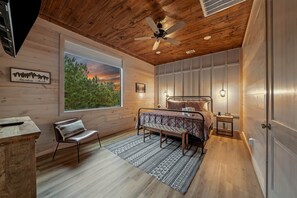 Un dormitorio con cama, una silla, un escritorio, ventilador de techo, ventana con vistas y una puerta.