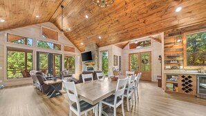 A spacious room with a wooden ceiling, large windows, a dining table with chairs, a sofa, and a kitchen area with a TV. - Hochatown Cabin Private Pool Hot Tub Game Room (Hochatown)