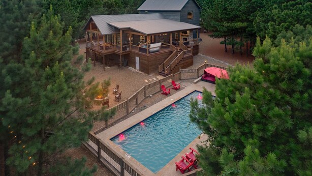 A two-story cabin with a deck, a swimming pool, and a forested backdrop. - Hochatown Cabin Private Pool Hot Tub Game Room (Hochatown)
