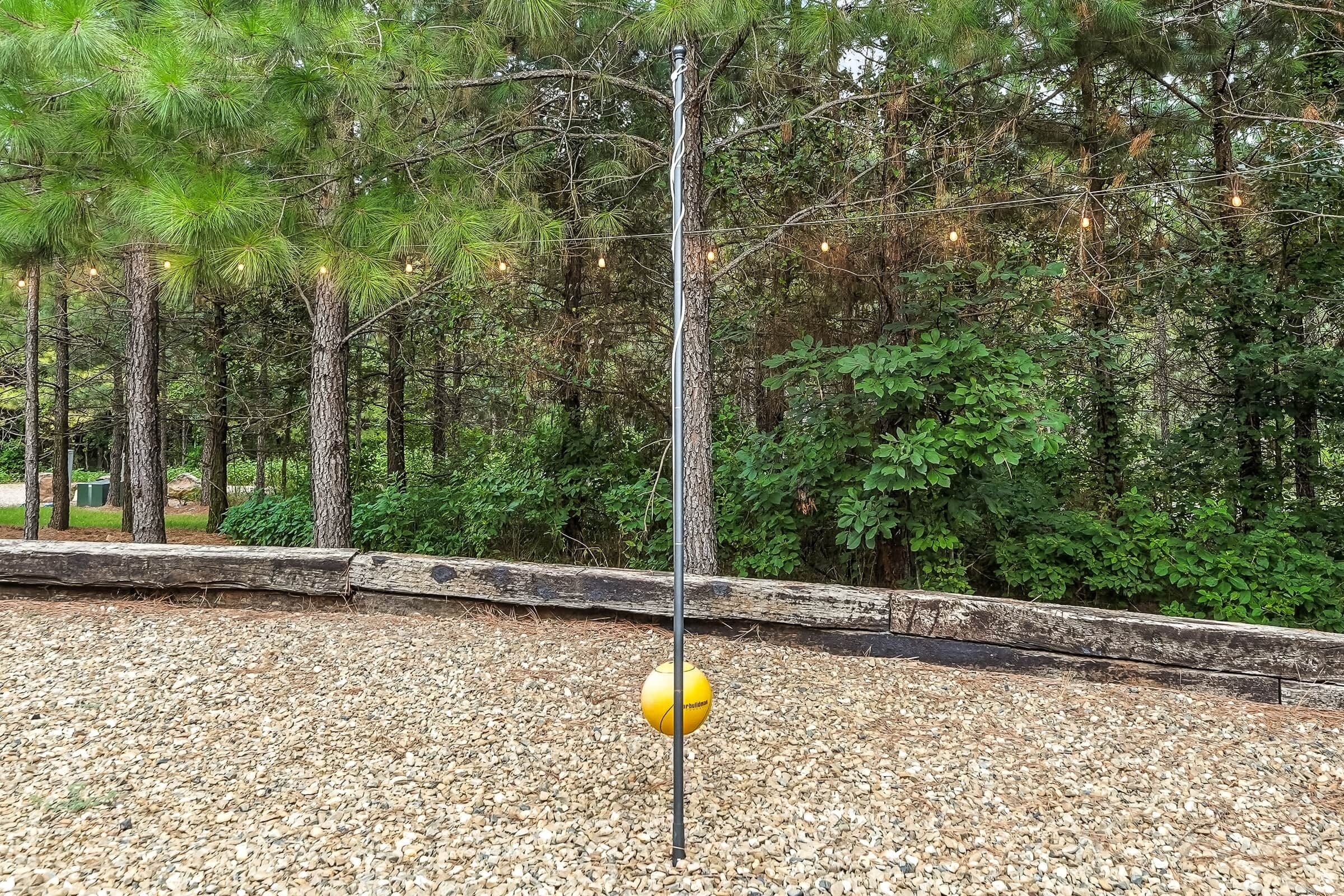 Un área con grava y una pelota amarilla en un poste, rodeada de árboles y un tronco de madera.