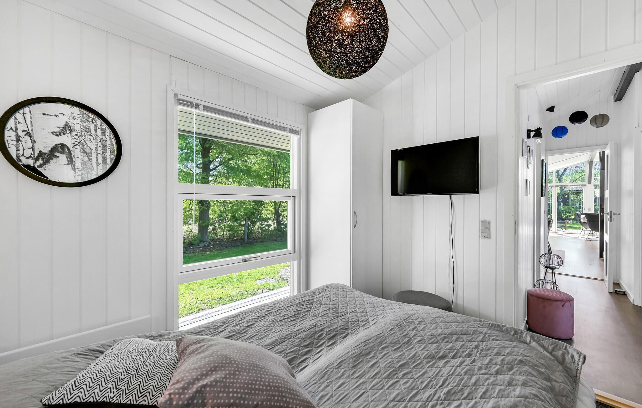 Ein Schlafzimmer mit einem Bett, einem an der Wand befestigten Fernseher und einem Fenster mit Blick ins Grüne.