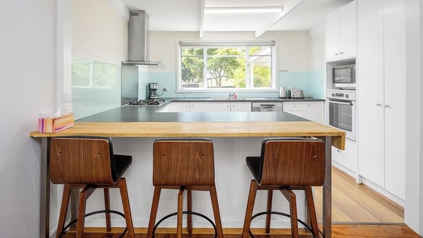 A modern kitchen with a central island, bar stools, a stove, and a window with a view of greenery. - Spacious Multifunctional 5BR Retreat with Backyard (Bayswater)