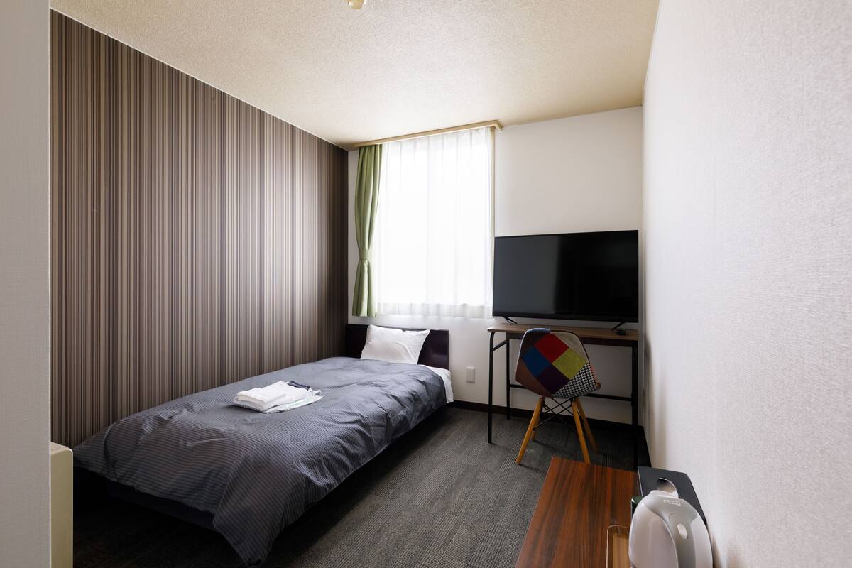 A hotel room with a bed, a television, a desk with a chair, and a window with curtains.