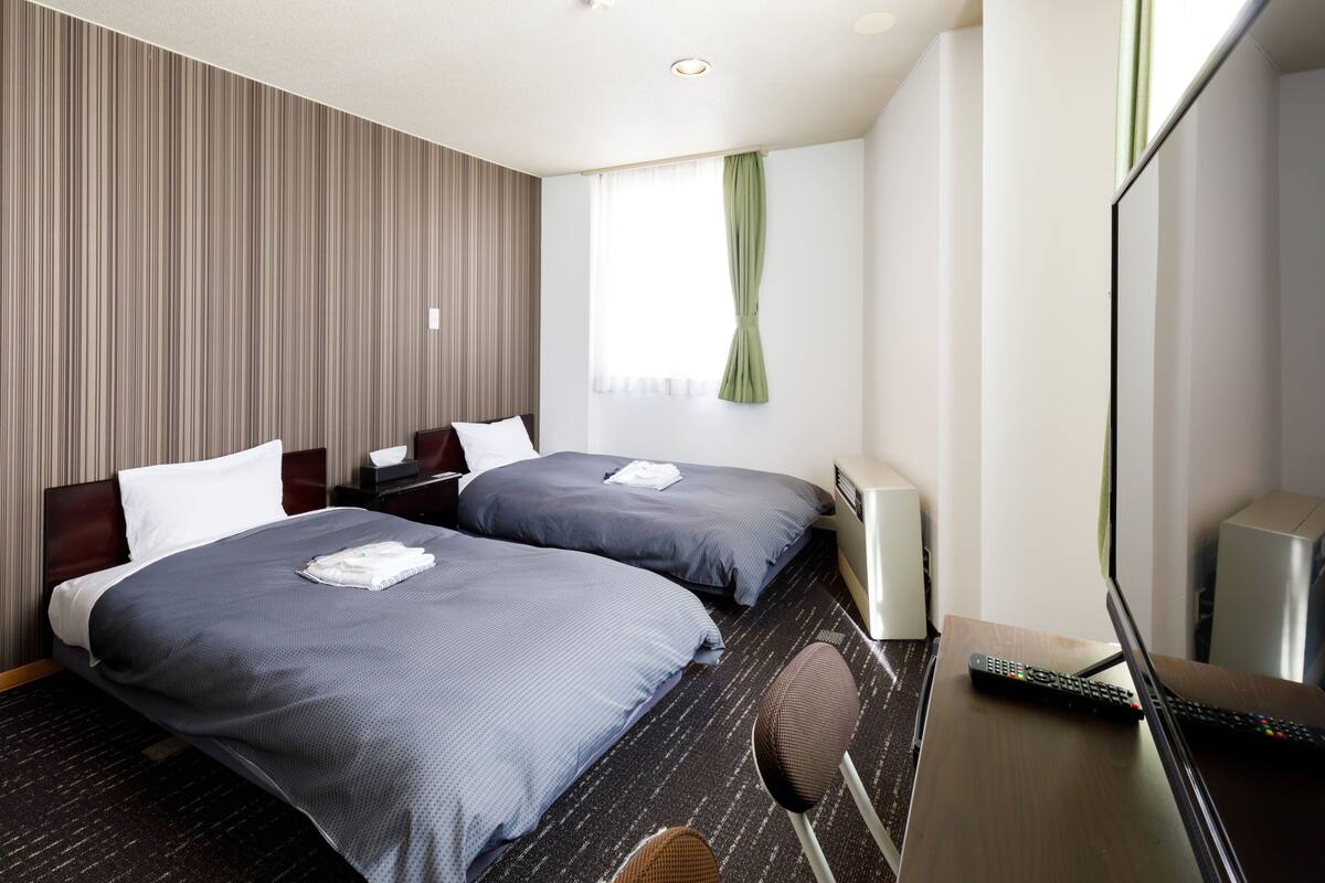 A hotel room with two beds, a desk with a computer and a remote control, a TV, and a window with curtains.
