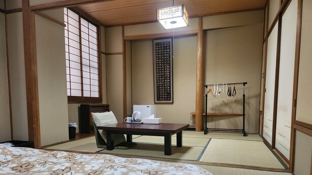 A traditional Japanese room with tatami flooring, a bed with a floral-patterned bedspread, a desk with a kettle and tissues, and a shoji screen.