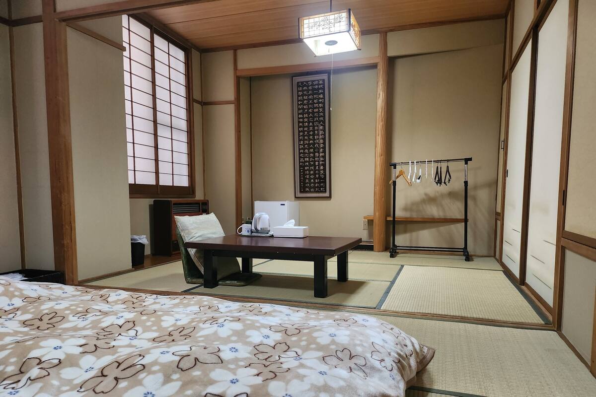 A traditional Japanese room with tatami flooring, a bed with a floral-patterned bedspread, a desk with a kettle and tissues, and a shoji screen.