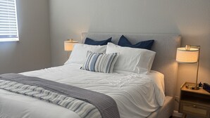 Een net opgemaakt bed met een grijze hoofdbord, witte lakens en een gestreept plaid. Twee nachtkastjes met lampen en een raam met zonwering.