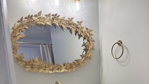Een badkamer met een witte wastafel, gouden kraanwerk en een spiegel met gouden lijst. Boven de spiegel hangen drie wandlampen.