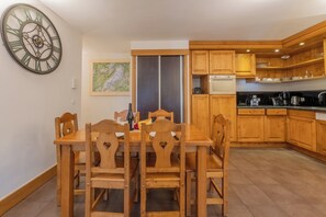 A dining area with a wooden table and chairs, a large wall clock, and a kitchen with wooden cabinets and appliances. - V Balme 12: Beside Ski Lift; Pool, Sauna & Gym, Vallorcine (Argentiere), France (Vallorcine (Argentiere))
