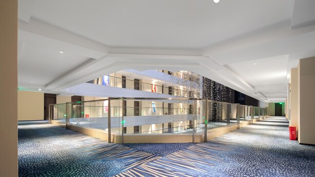 Eine geräumige Hotelhalle mit einer zentralen Glasvitrine, einem gemusterten Teppich und einer hohen Decke.