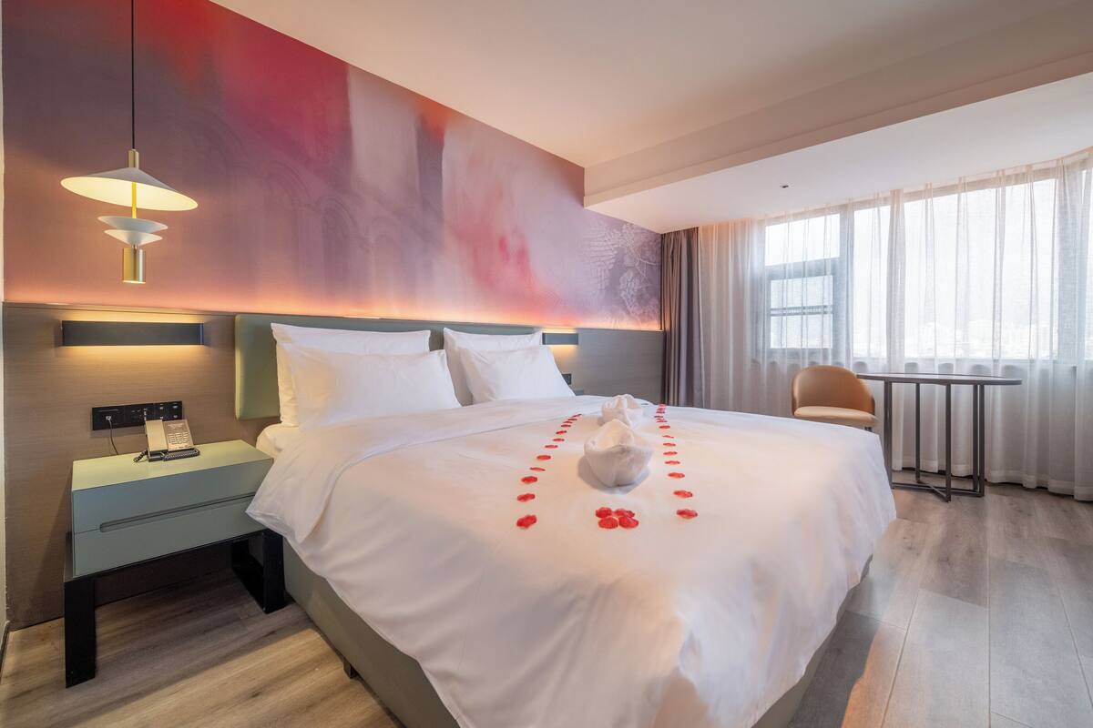 Habitación de hotel con una cama grande, una mesita de noche con teléfono y control remoto, una silla y una ventana con cortinas traslúcidas.
