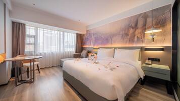 Habitació d'hotel amb un llit gran, una taula de nit, un escriptori amb cadira i una finestra ampla amb cortines.