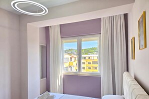 Habitació d'hotel amb un llit, una finestra amb cortines, vistes a edificis i una làmpada circular al sostre.