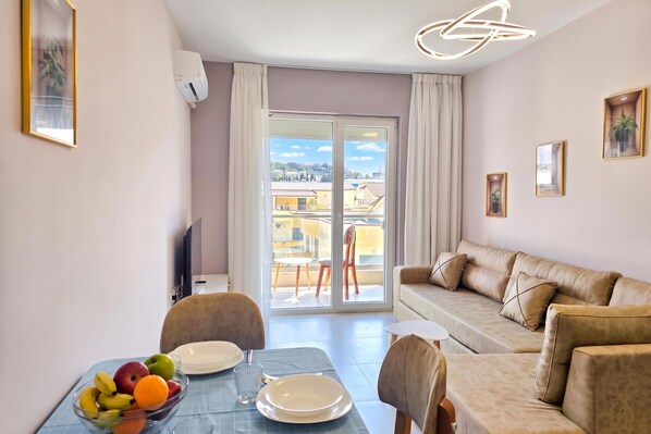 Un saló modern amb un sofà, taula de dinar amb plats i fruita, i una vista per una porta de vidre.