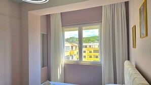 Una habitación de hotel con cama, ventana con cortinas y vistas a edificios, además de una lámpara con forma circular.