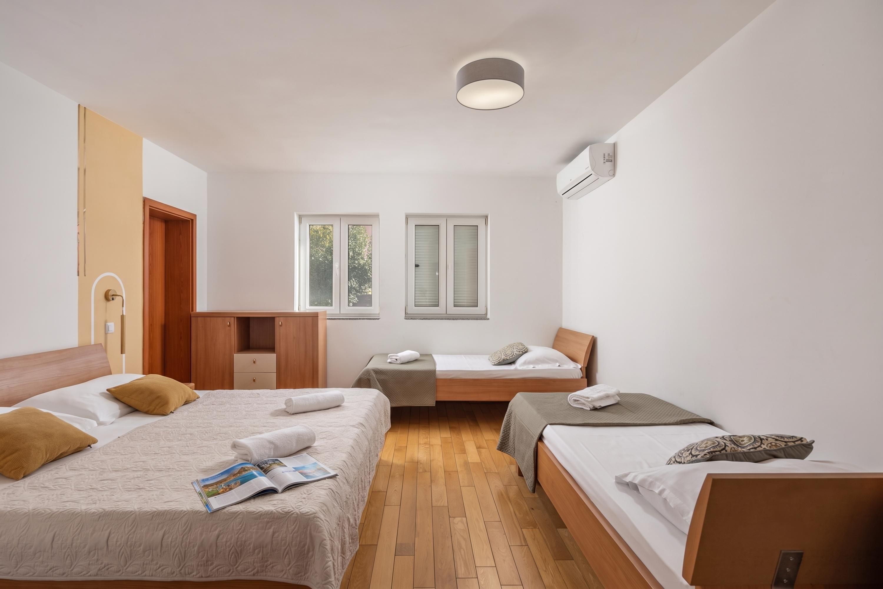 Hotellihuone, jossa on kaksi erillistä sänkyä, puinen vaatekaappi, pieni työpöytä ja ikkuna, jossa on sälekaihtimet.